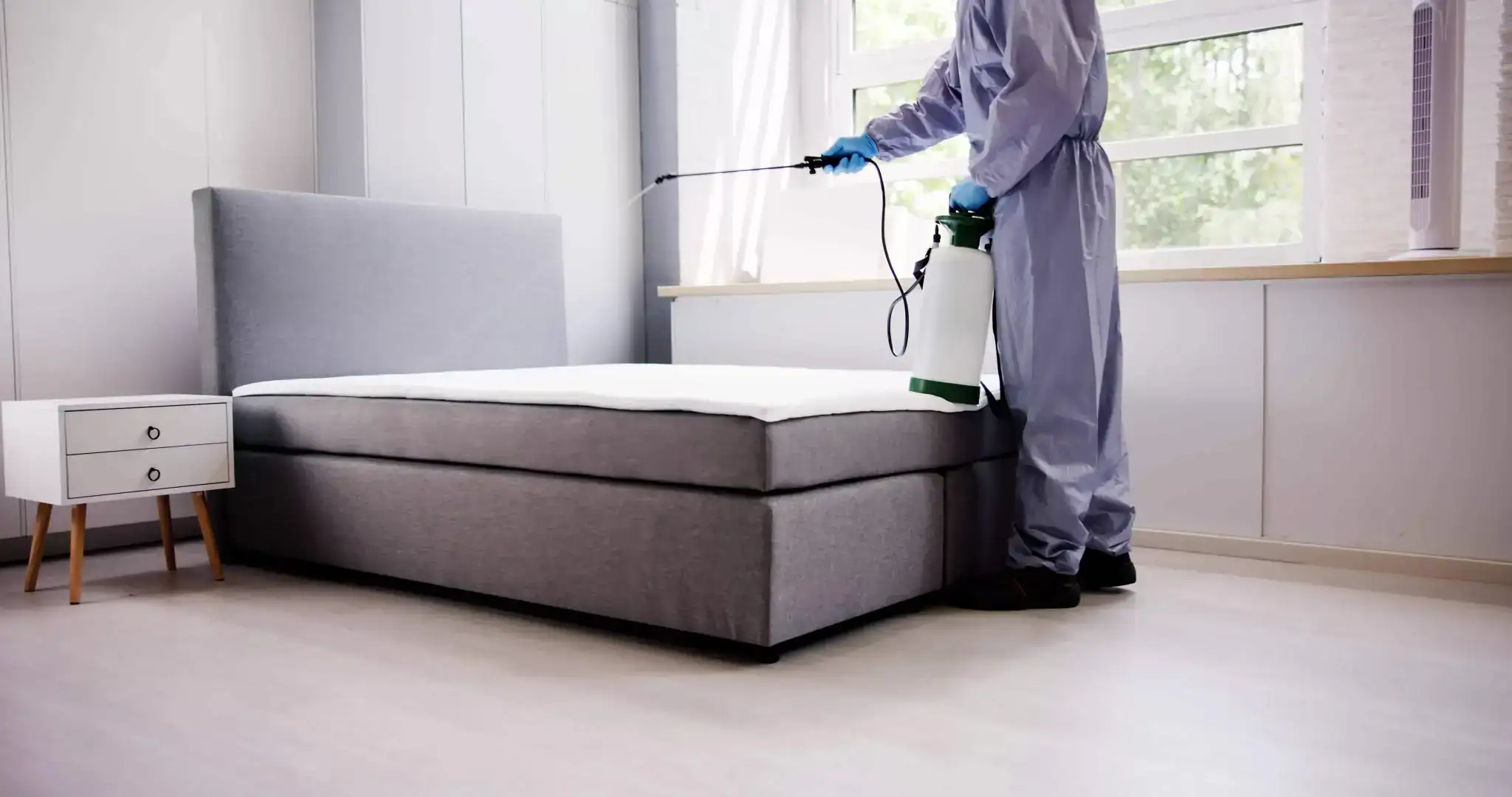 A person in protective clothing and gloves sprays disinfectant on a bed in a bright, modern bedroom with a large window and minimal furniture, demonstrating expert pest control Genesee County, MI services.
