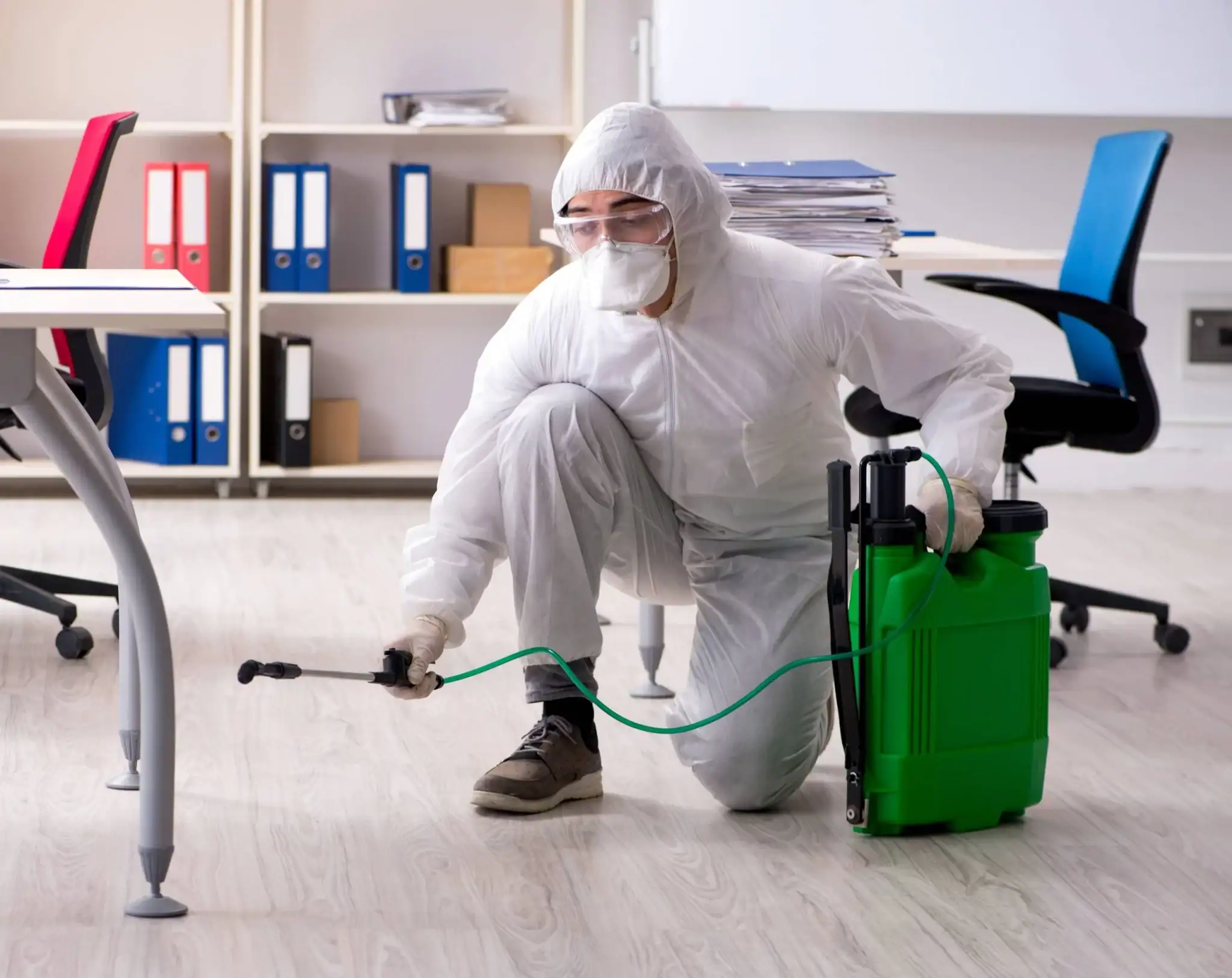 A person wearing a white protective suit, mask, and gloves is kneeling on the floor, using a green sprayer to disinfect an office space in Genesee County, MI. Pest Control Genesee County services help ensure clean and safe work environments.