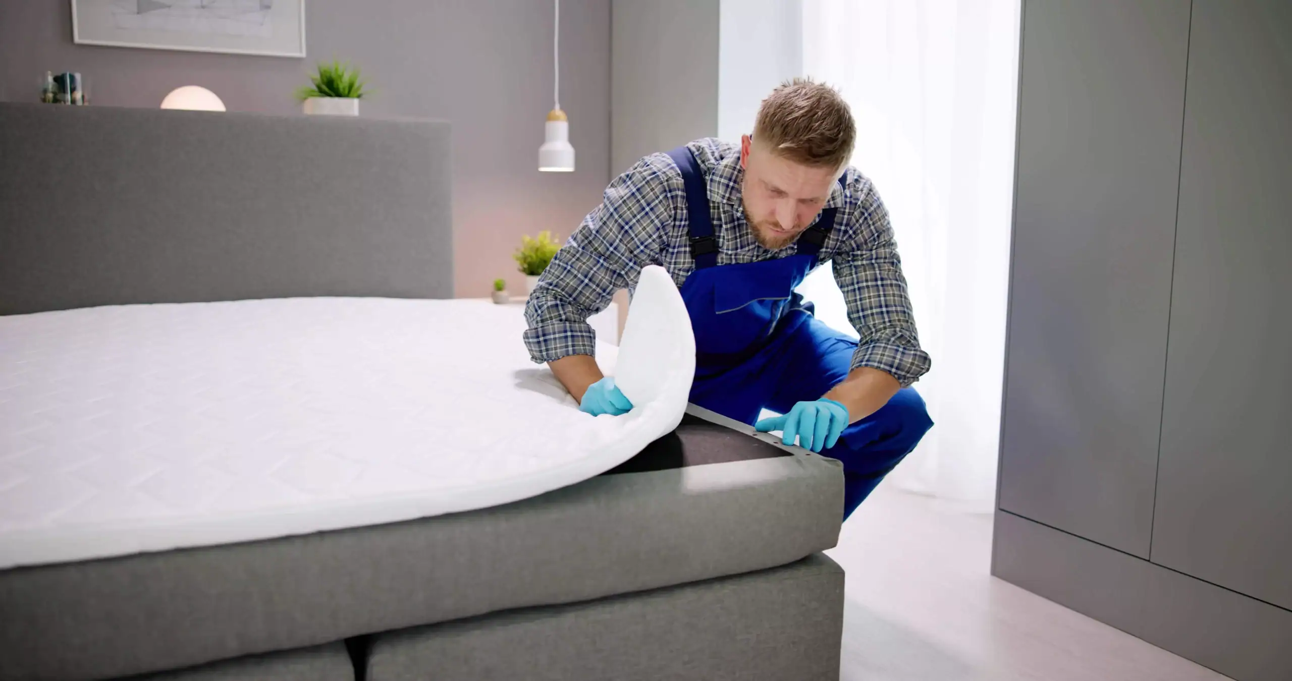 A man in blue overalls and gloves inspects the corner of a mattress on a grey bed in a bright, modern MI bedroom with plants and pendant lighting, showcasing expert Pest Control Genesee County services.