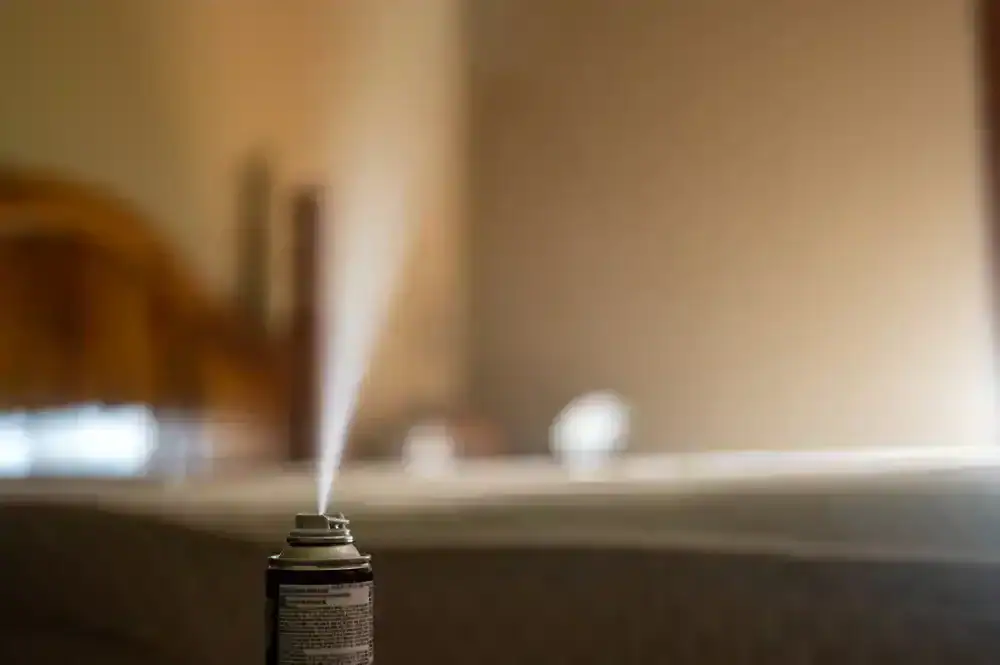 A close-up of an aerosol spray can releasing a fine mist into a softly lit room in MI, with a blurred background, suggesting use of air freshener, disinfectant, or pest control methods common in Genesee County.